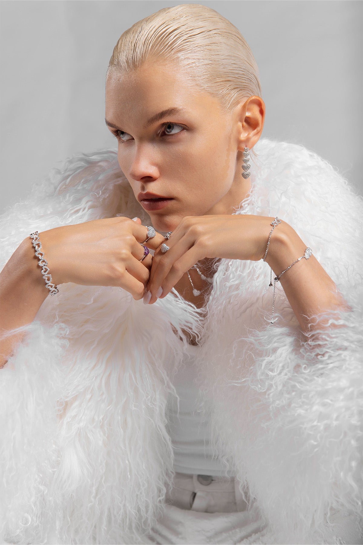 Model with APM MONACO heart-shaped jewelry and wearing a white feathered coat against a gray background