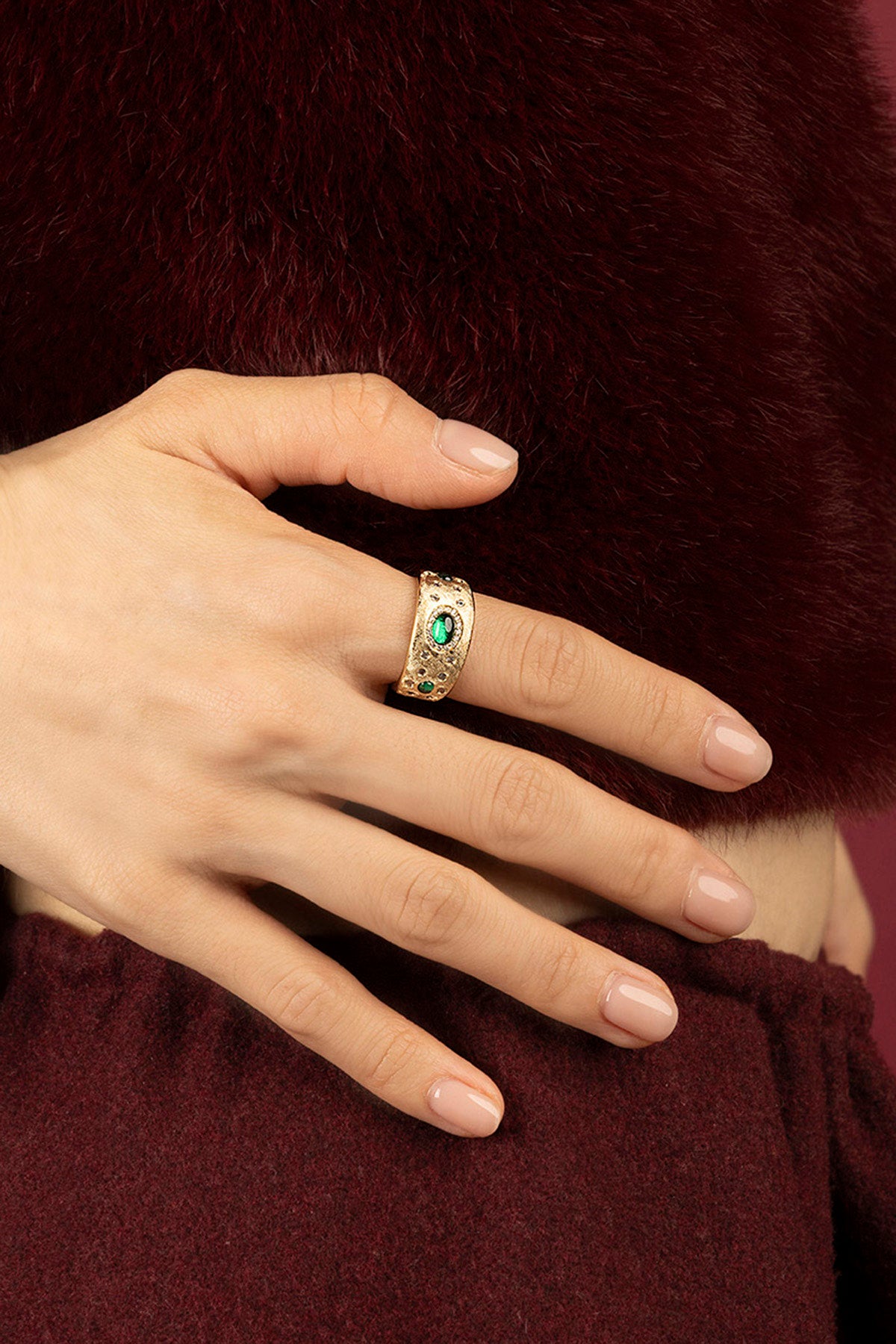Woman wearing APM Ring, a golden statement with emerald allure against burgundy warmth.