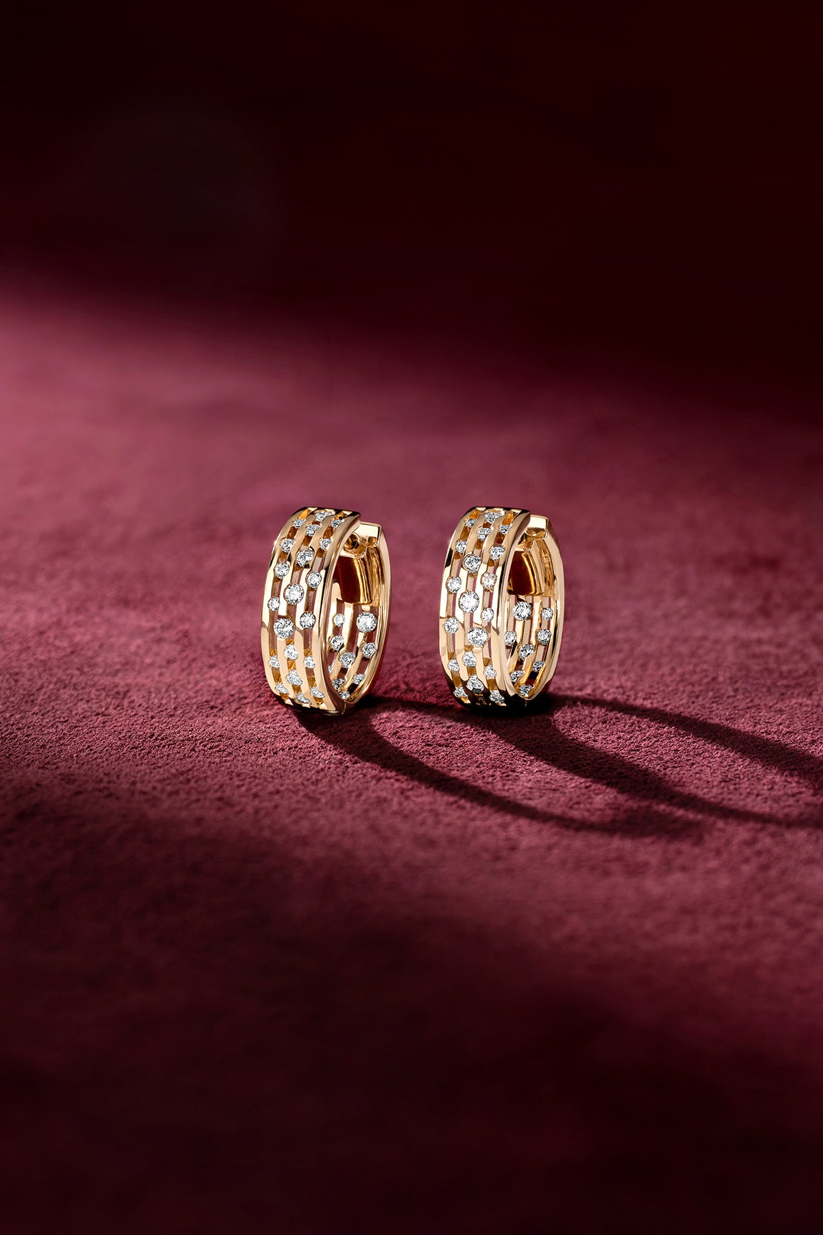 APM Hoop Earrings, golden architecture sparkling softly against a velvet crimson glow.