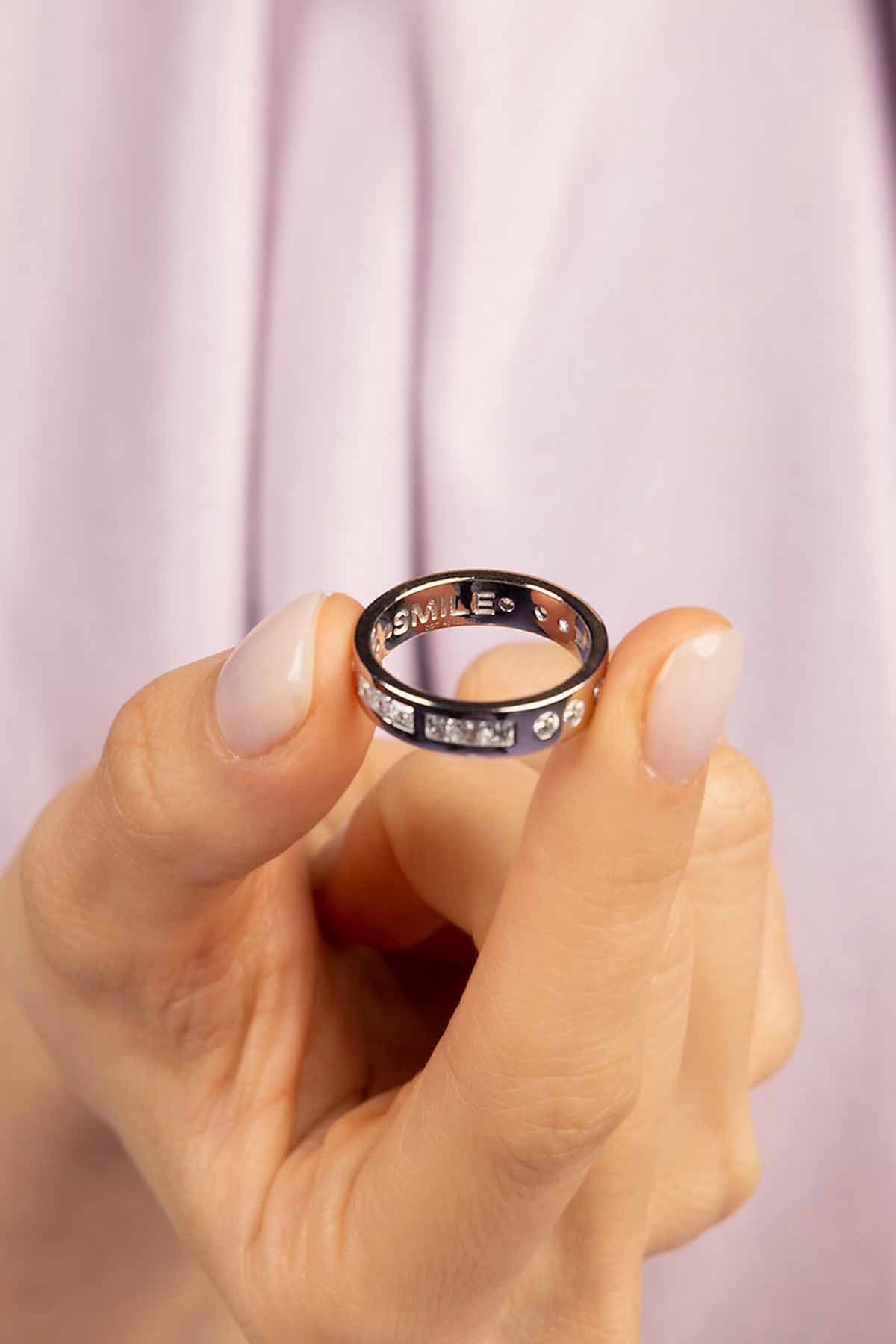 Hand holding a silver ring with 'SMILES' engraving against a pink background