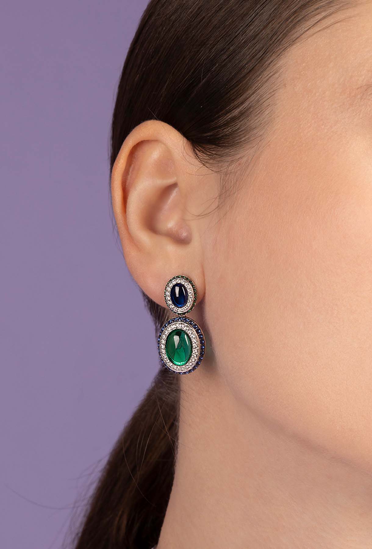 Close-up of a person wearing APM MONACO silver earrings with bold green and blue stones against a purple background