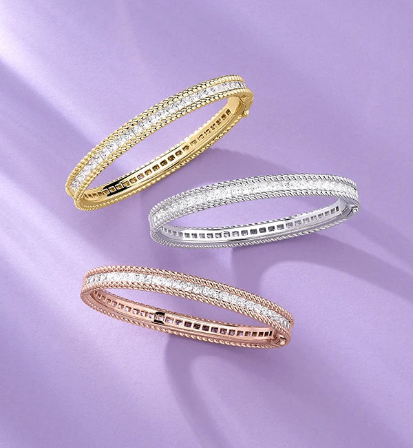 Three APM MONACO torsade bangles in yellow gold, silver, and rose gold on a purple background